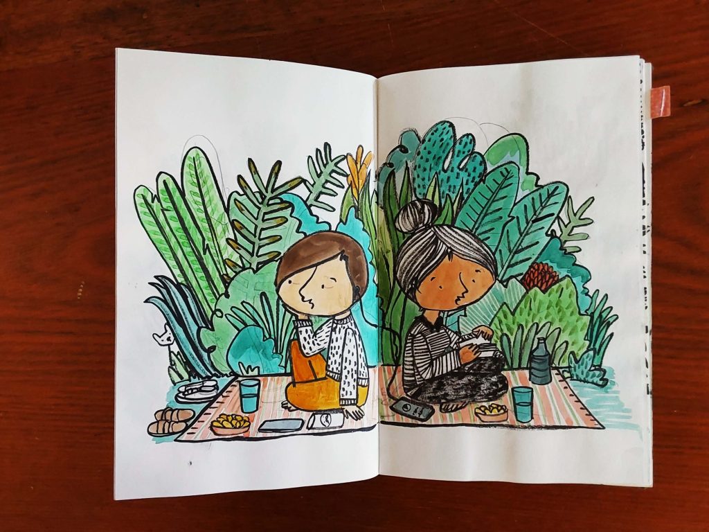 Caderno aberto com um desenho de uma dupla de costas um pro outro fazendo piquenique sobre uma toalha com comidas e bebidas e plantas ao fundo.