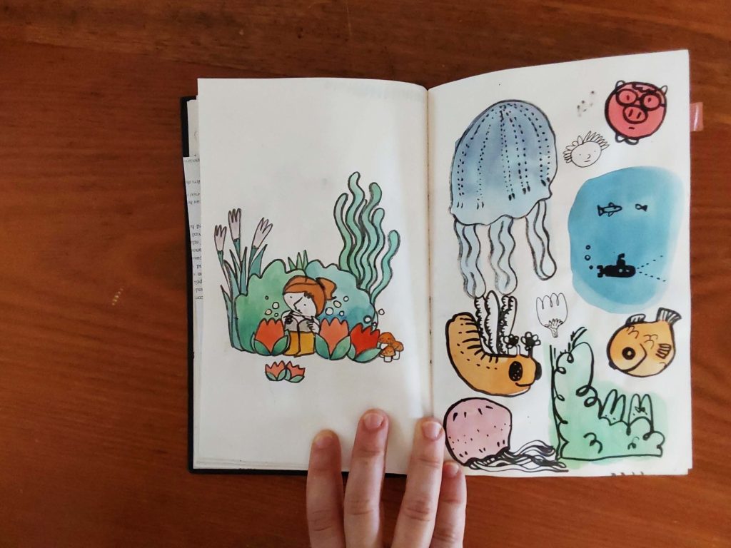 Foto de um caderno aberto com uma lapiseira e uma borracha do lado esquerdo. Na página esquerda do caderno se vê um desenho de uma menina sentada junto a uma moita de plantas com três flores vermelhas gigantes, três cogumelos marrons e duas flores menores na frente. Na outra página se vê um animal parecido com uma lagarta marrom com asas finas, uma água viva azul, um submarino negro em um fundo azul, um peixe, uma água viva rosa, uma planta e um porquinho rosa