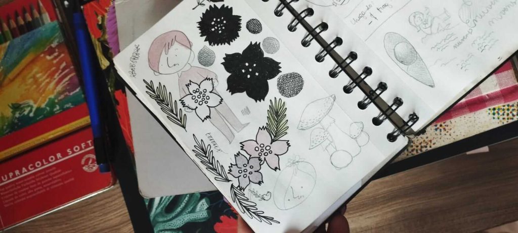 foto de um caderno aberto com deenhos de sakuras negras e rosadas, além de folhas verdes, e um menino segurando uma sakura branca. Além disso, a página tem folhas verdes pinadas e cogumelos desenhados a lápis. Abaixo do caderno se vê uma pilha de outros cadernos, uma lapiseira e uma caixa de lápis de cor.