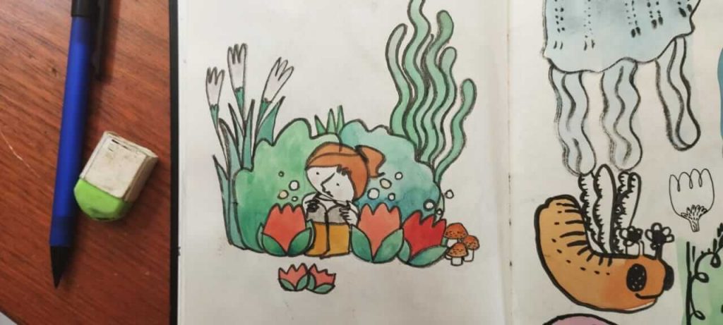 Foto de um caderno aberto com uma lapiseira e uma borracha do lado esquerdo. Na página esquerda do caderno se vê um desenho de uma menina sentada junto a uma moita de plantas com três flores vermelhas gigantes, três cogumelos marrons e duas flores menores na frente. Na outra página se vê um animal parecido com uma lagarta marrom com asas finas e pedaços de outros desenhos, como uma água viva, no canto direito superior